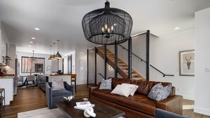 A modern living room with a brown leather sofa, a coffee table, and a large black pendant light. A staircase with a wooden finish and black railings is visible in the background. - Luxury Townhouse with Rooftop Deck & Hot Tub, Close to Skiing - Skyline Retreat (Steamboat Springs)