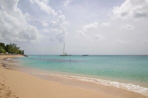 A beach with clear turquoise water, a sailboat anchored in the distance, and a sandy shore with footprints. - Large pool, gardens and close to beach - Aqua Bliss (Sunset Crest)