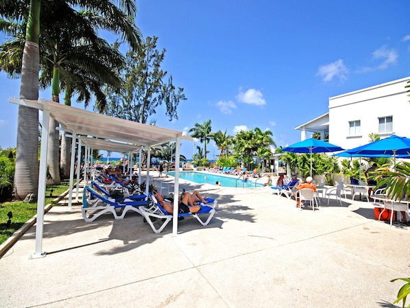 A poolside area with lounge chairs and umbrellas, surrounded by palm trees and a clear sky.
