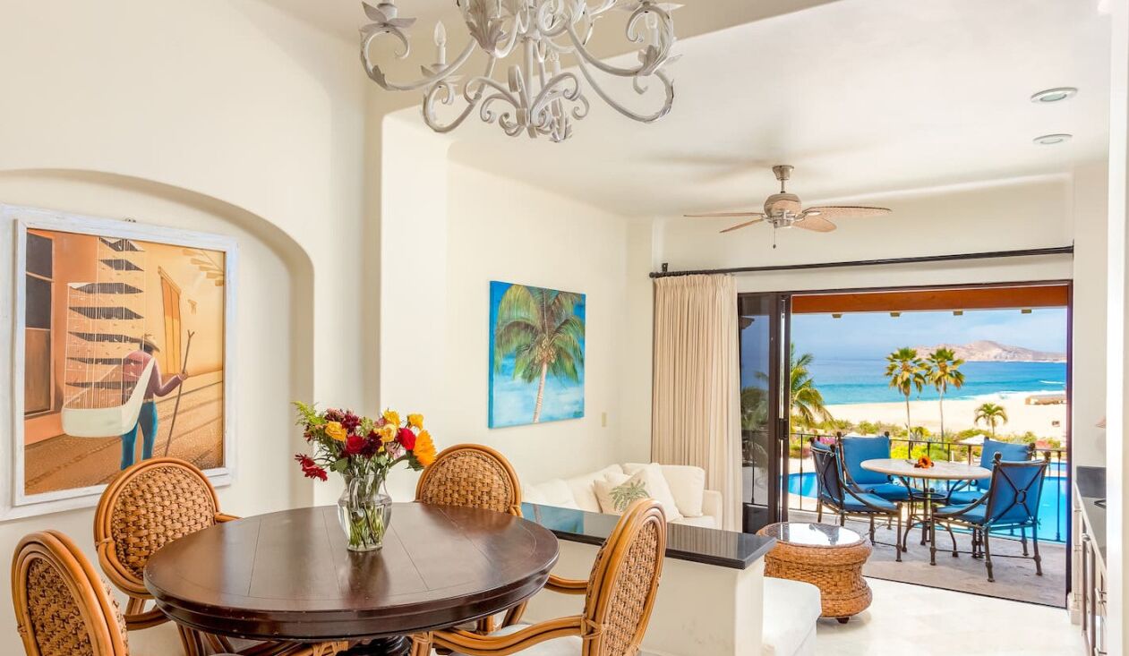 A dining area with a round table, wicker chairs, and a vase of flowers. There is a view of a beach and ocean through a large glass door.