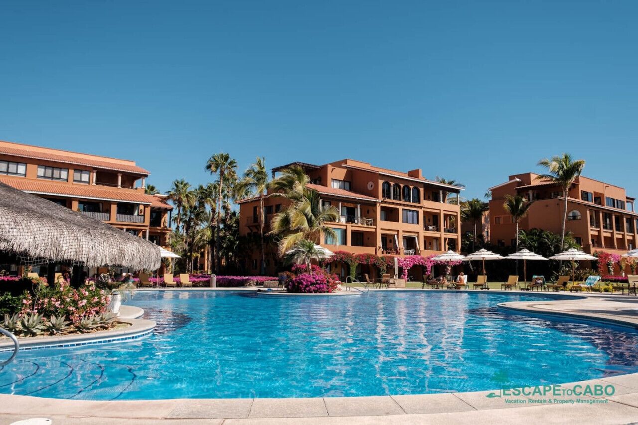 A resort with a large swimming pool, palm trees, and a thatched-roof structure.