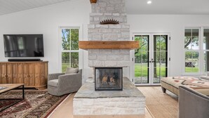 A modern living room with a stone fireplace, wooden beams, a television, and a large window.
