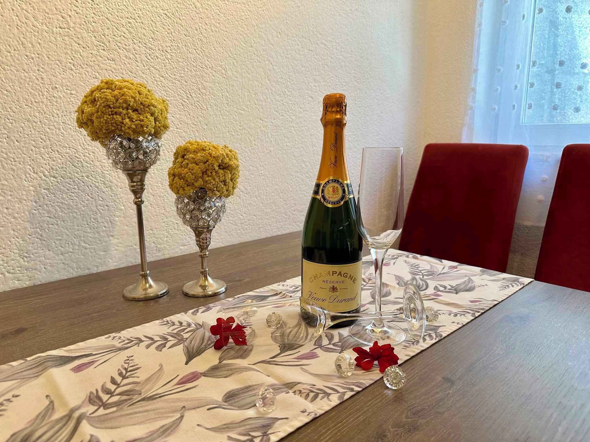 Une table dressée avec une bouteille de champagne, deux vases en cristal ornés de fleurs jaunes et un verre posé sur une nappe à motifs floraux.