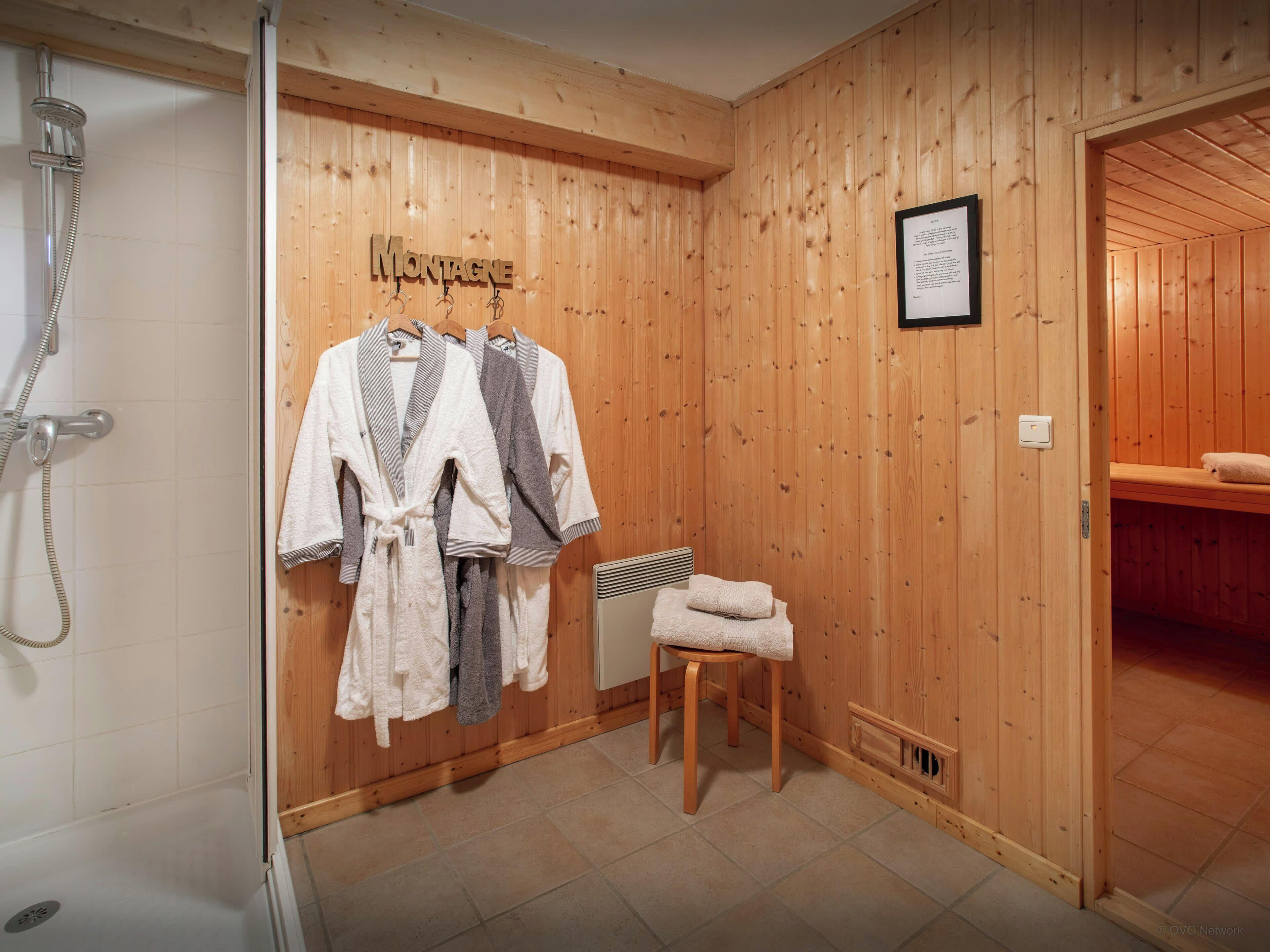 A wooden-paneled room with a shower, a bench, and two robes hanging on a rack.
