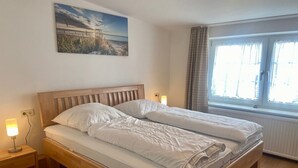 A double bed with a wooden headboard, two bedside tables with lamps, a window with curtains, and a painting on the wall. - 5060 Saarstr. 8a, Deutschland (Dahme)