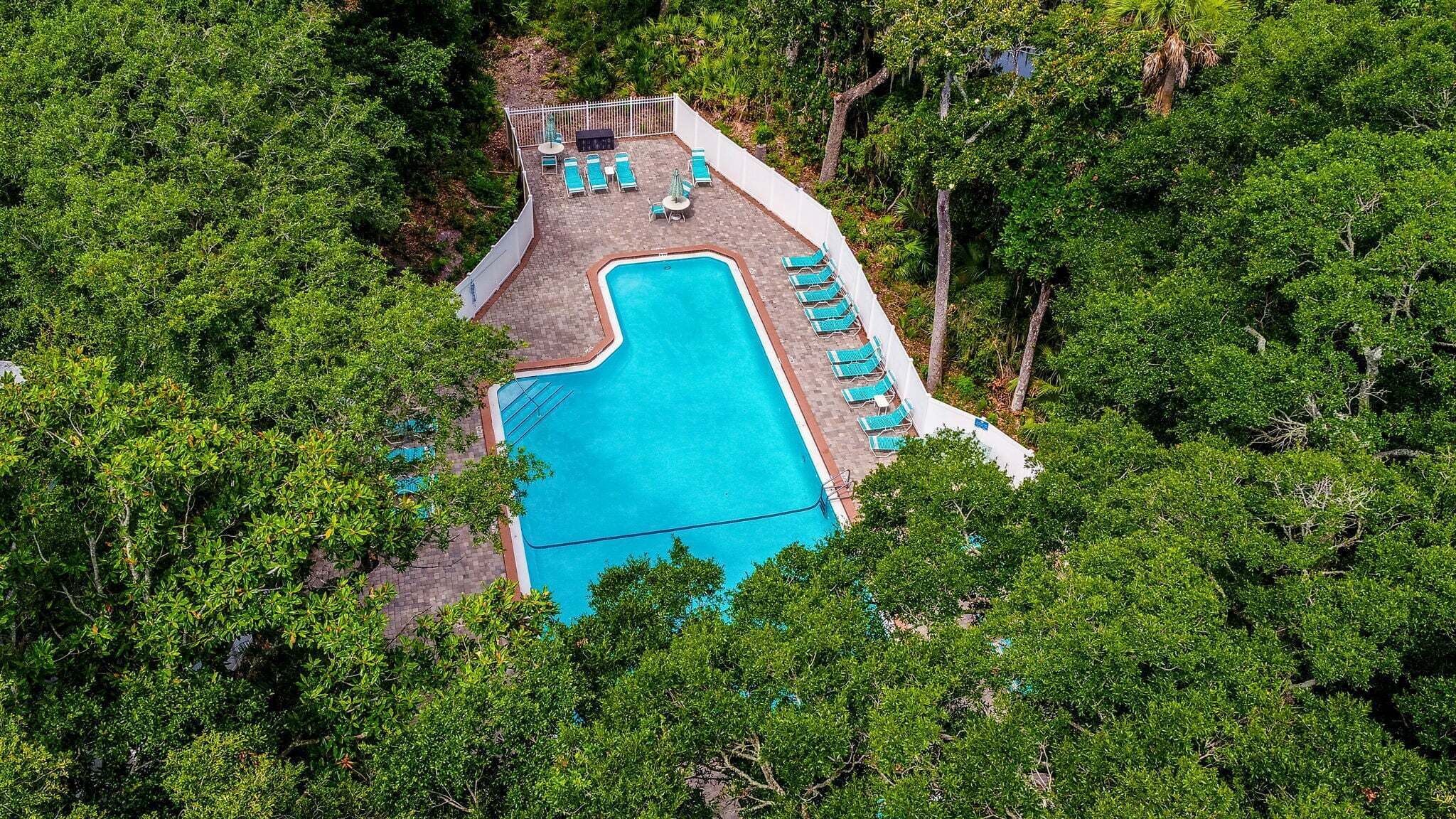 Une vue aérienne d’une piscine entourée d’une végétation luxuriante, avec des chaises longues et une terrasse délimitée par une clôture.