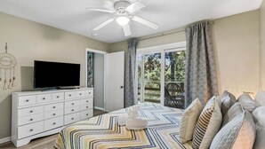 Une chambre à coucher avec un lit, un téléviseur posé sur une commode, un ventilateur de plafond et une vue sur l’extérieur.