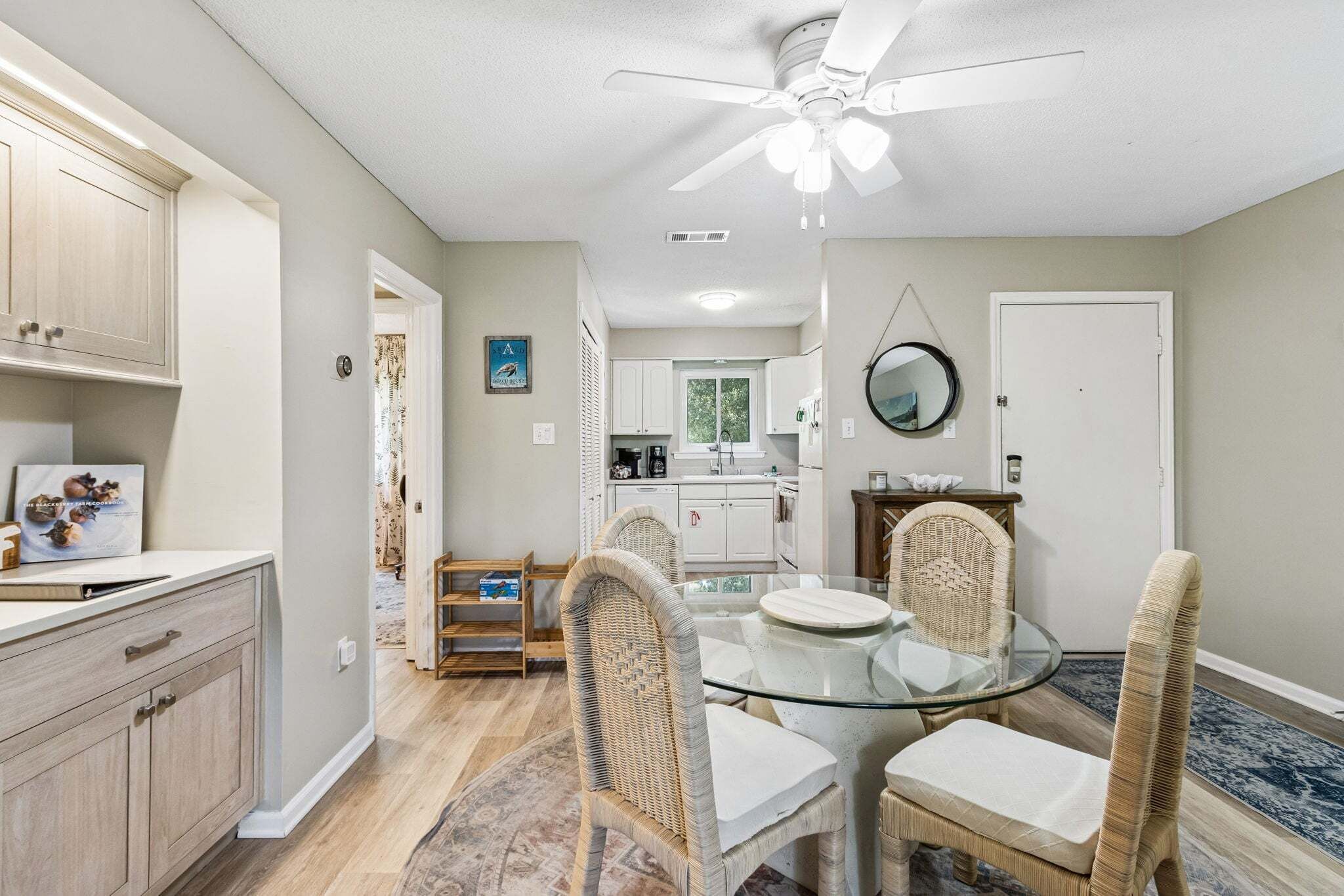 A dining area with a glass table and wicker chairs, a ceiling fan, and a kitchen in the background.