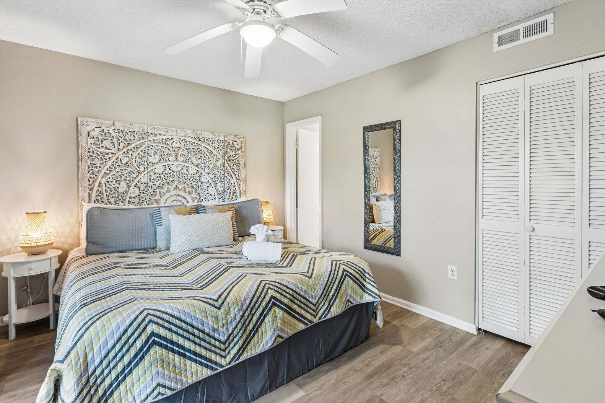 A bedroom with a bed, a ceiling fan, a nightstand, a bedside lamp, a mirror, and a closet.