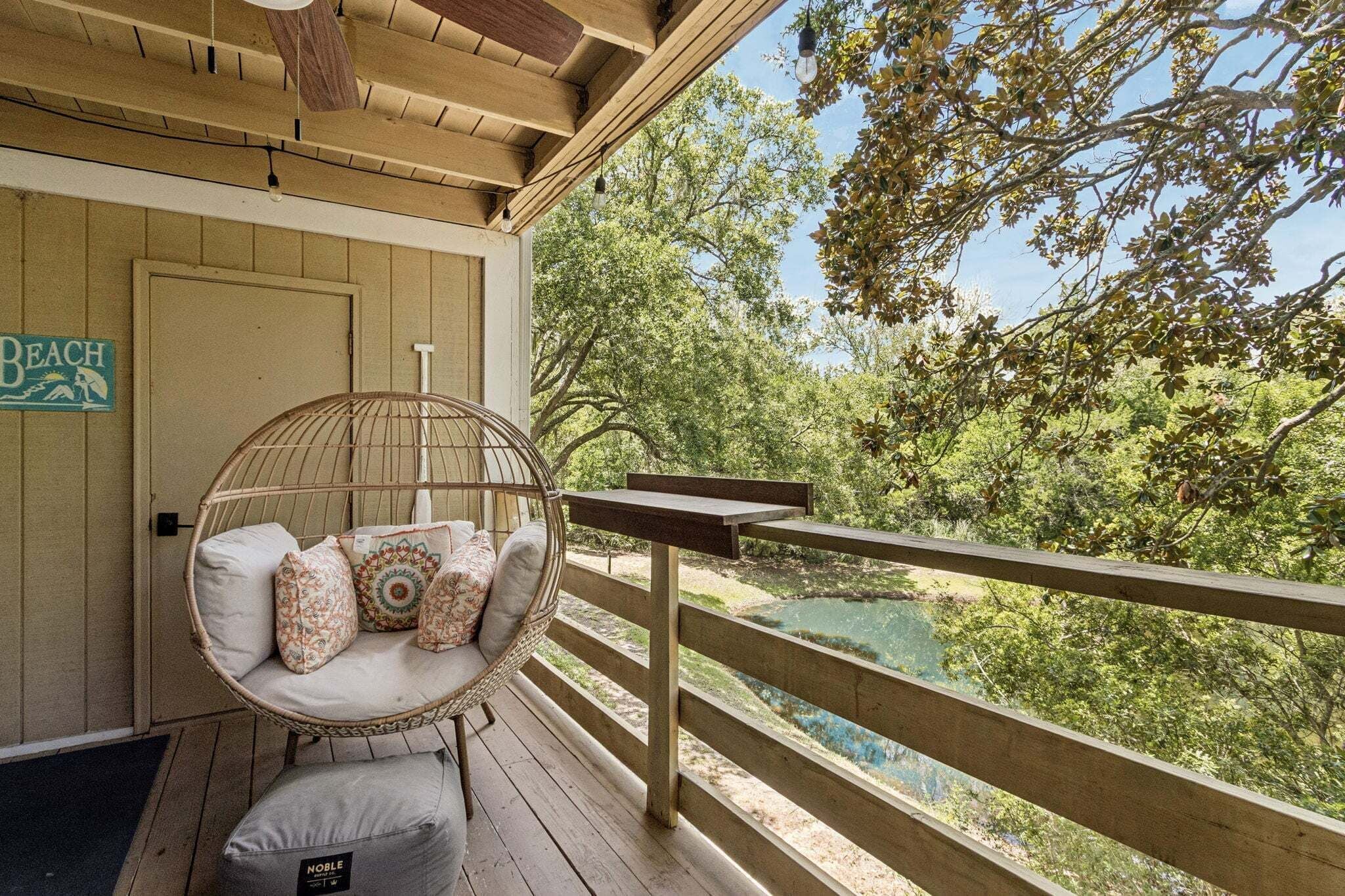 A wooden deck with a wicker chair and cushions, overlooking a natural landscape with trees and a body of water.