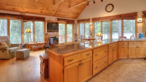 Un interior de cabaña espacioso con pisos de madera, una cocina grande con armarios de madera, una zona de comedor con una mesa y sillas, y ventanas amplias que ofrecen vistas al bosque.