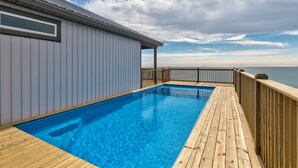 A wooden deck with a swimming pool, a covered area with a window, and a view of the sea. - Salty Piece of Land | Nightly openings 8/22. Pool. (Dauphin Island)