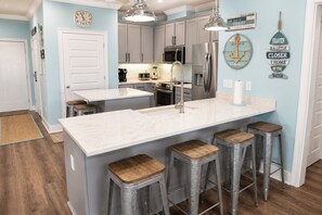 A kitchen with a large island, bar stools, stainless steel appliances, and a clock on the wall. - Lighthouse View | Gather by the Gulf, book your xmas stay with us (Dauphin Island)