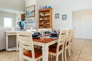 Un comedor con una mesa de madera dispuesta para cuatro personas, sillas blancas y una estantería con objetos decorativos.