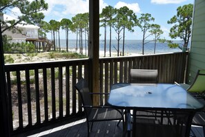 Une terrasse en bois avec une table et des chaises en verre, donnant sur une plage bordée de pins et sur laquelle on aperçoit un bâtiment au loin.