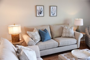 A living room with a beige sofa, two framed wall art pieces, a side table with a lamp, and a patterned ottoman.