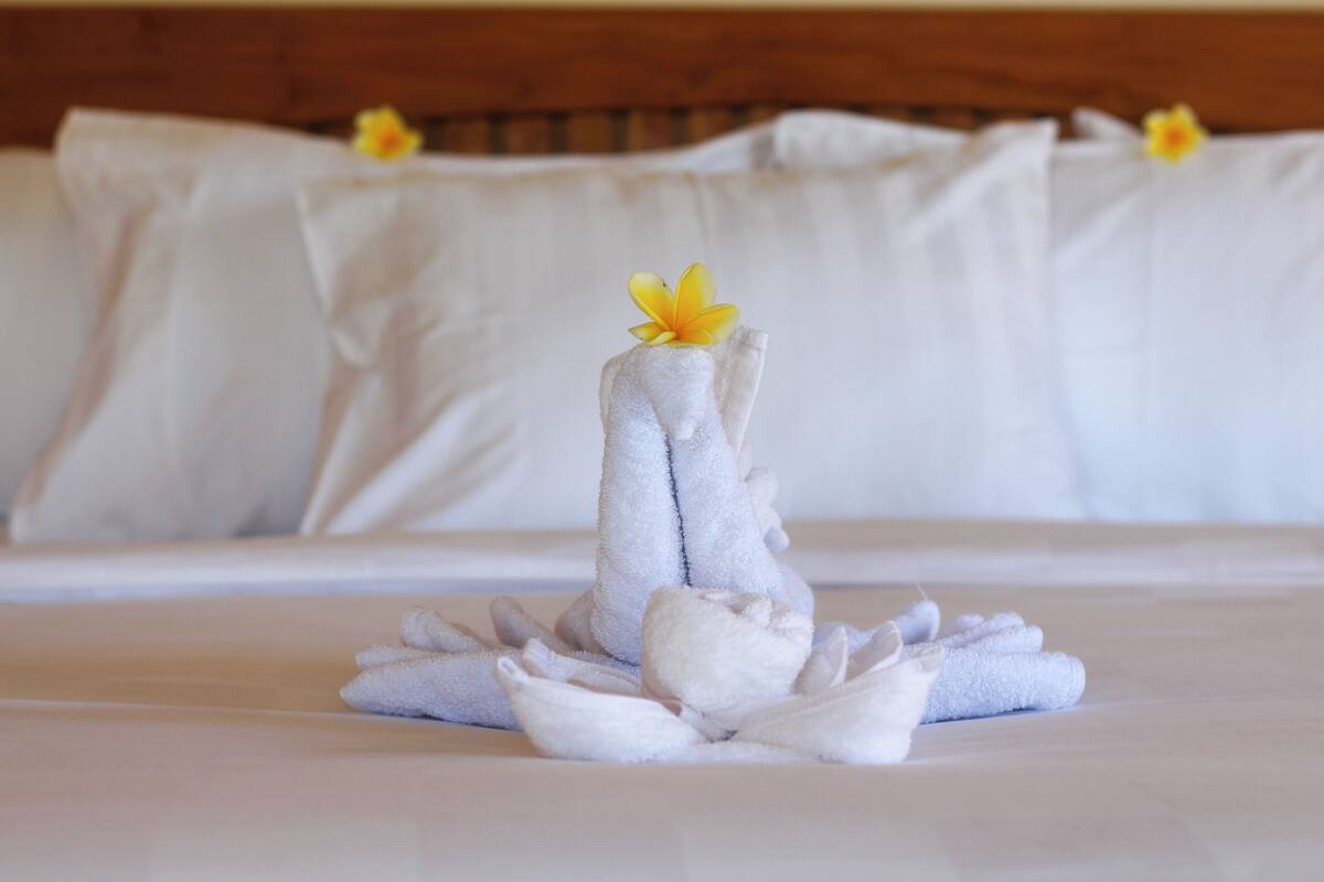 A neatly arranged towel sculpture with a flower on a bed, set against a backdrop of white pillows and a wooden headboard.