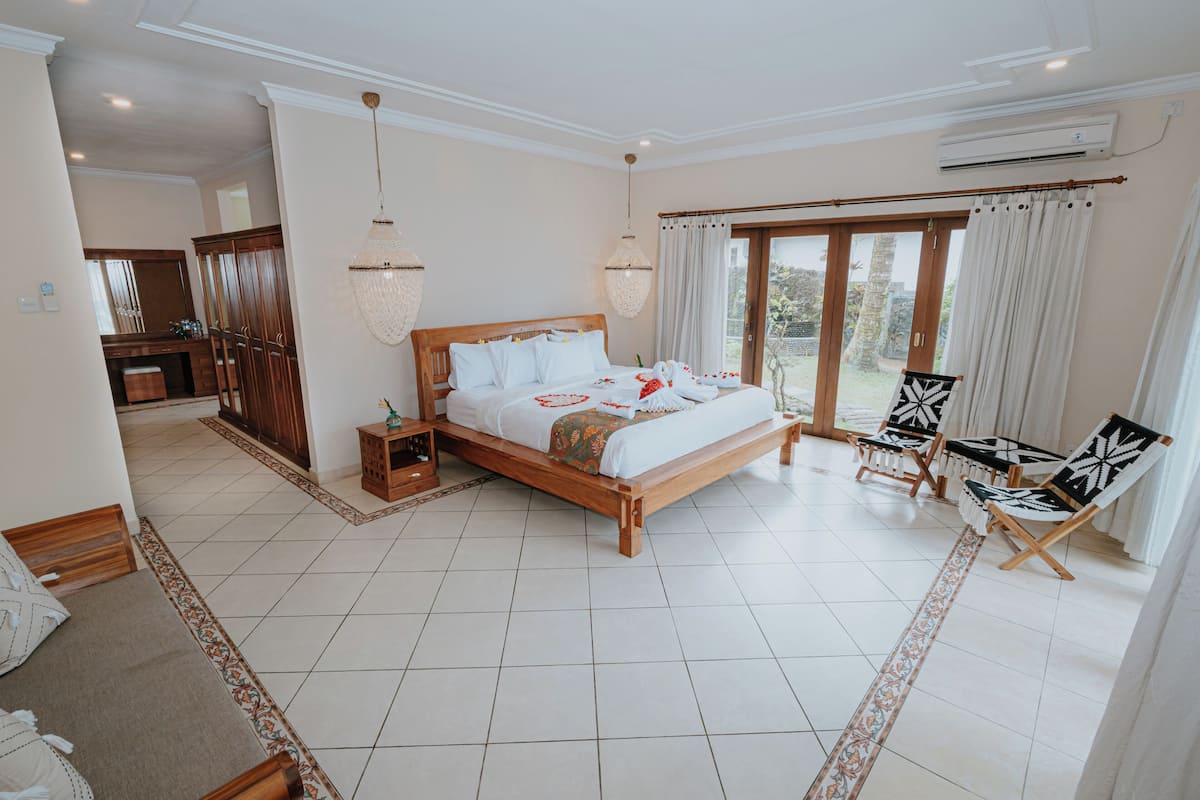 A spacious bedroom with a large bed, a sitting area with chairs, and a view through the glass door.