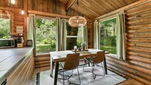 Interior rústico de cabana com mesa de jantar de madeira, duas cadeiras, luminária pendente e amplas janelas com vista para a floresta.