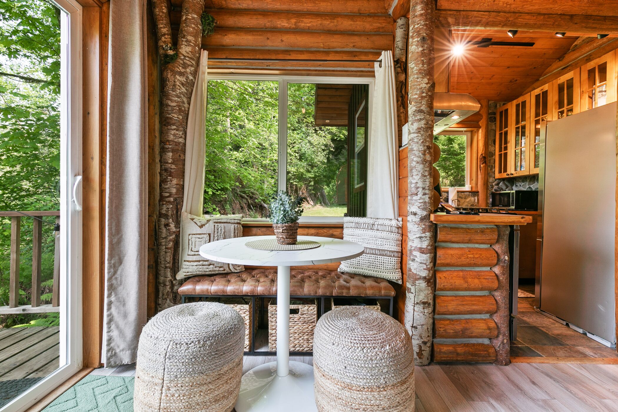 L’intérieur d’un chalet rustique, avec un banc en bois, une table ronde et une vue sur la forêt par une grande fenêtre.
