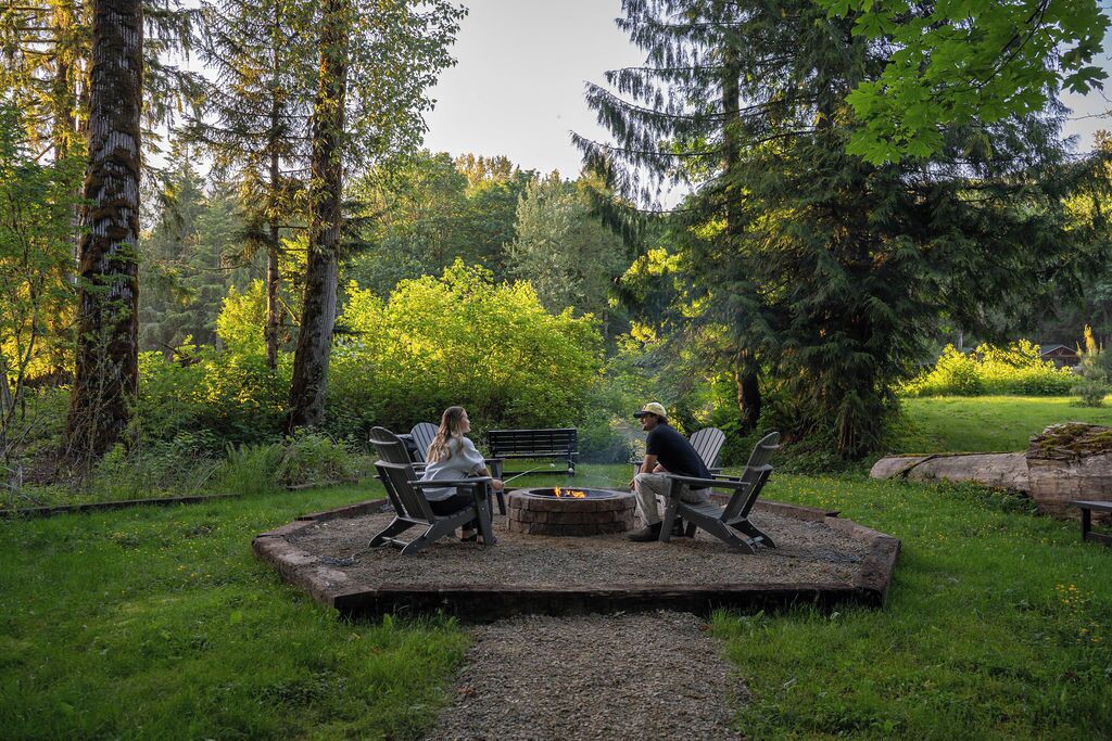 Un espacio con una fogata, dos sillas Adirondack y un banco, rodeado de un paisaje boscoso.