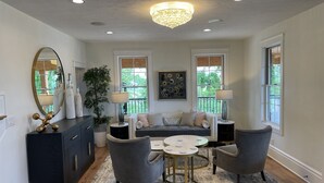 A modern living room with a grey sofa, two armchairs, a coffee table, and a chandelier. There are two windows with views of greenery, a large mirror, and a dark wooden cabinet. - 7 Bedrooms, 2 Houses all for 1 Price, 5 BR Large House & 2 BR Carriage House (Morgantown)
