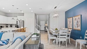 A modern kitchen with white cabinets, a dining area with a white table and chairs, and a living room with a gray sofa and coffee table.