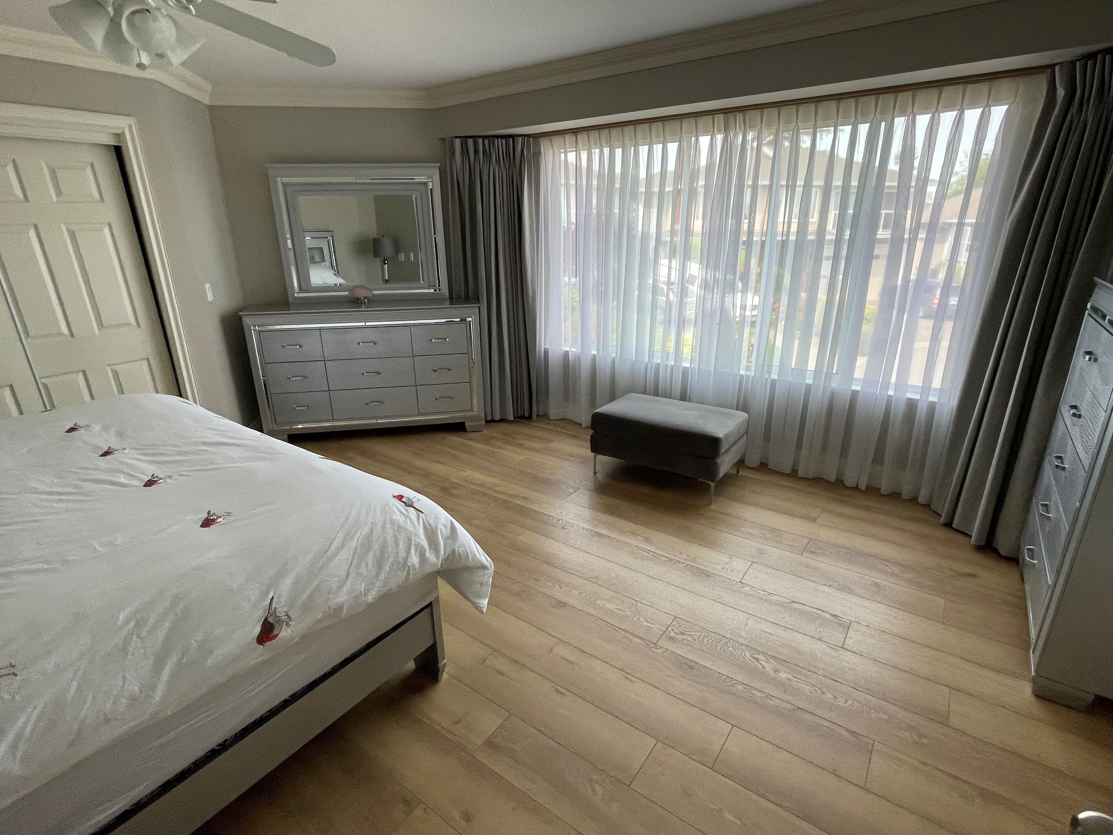 Un dormitorio con cama, cómoda, espejo, otoman y ventana con cortinas.