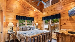 A wooden cabin room with a bed, two bedside lamps, a window with a view, and a ceiling fan. - Lovin' the View - Gorgeous views plus a true cabin feel! (Gatlinburg)