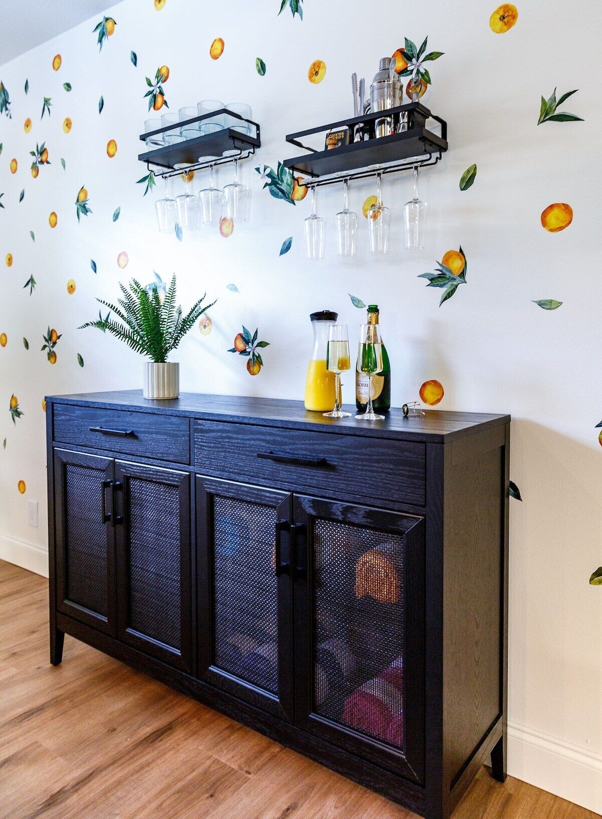 A dark blue cabinet with glass doors, a potted plant, and a shelf with glasses and bottles against a wall with fruit decals.
