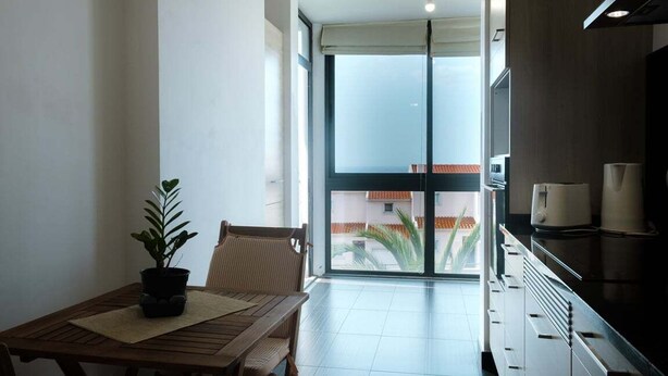 Ein Balkon mit Esstisch, Stühlen, einem Blumentopf und Blick auf Gebäude durch eine Glastür.