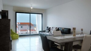Ein modernes Wohnzimmer mit Esstisch, grau-weißem Sofa und einem großen Fenster mit Blick auf Gebäude.
