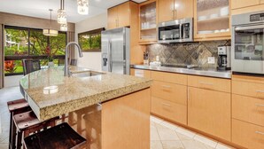 A modern kitchen with granite countertops, stainless steel appliances, wooden cabinets, and a large window offering a view of greenery. - Ground Floor Ease - Walk to Beach & Pool (Lahaina)