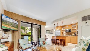 A modern living room with a glass coffee table, comfortable seating, and a kitchen area with wooden cabinets and a large island. - Ground Floor Ease - Walk to Beach & Pool (Lahaina)