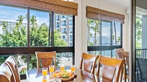 Un coin repas avec une table dressée pour quatre personnes, de grandes fenêtres offrant une vue sur les palmiers et les bâtiments, et un balcon avec des chaises.
