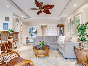 Ein geräumiges Wohnzimmer mit einem großen Deckenventilator, einem grauen Sofa, Korbstühlen, einem Glastisch und einer Topfpflanze.