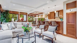 A spacious living area with a sofa, chairs, a coffee table, and a kitchen area with wooden cabinets and a countertop. - Modern Beach Condo with Platinum Rating (Lahaina)