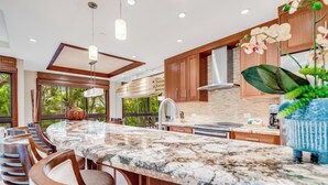 A modern kitchen with a large island, wooden cabinets, and a view of greenery outside. - Modern Beach Condo with Platinum Rating (Lahaina)