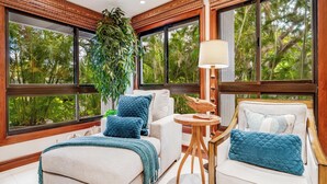 A modern room with large windows, a large plant, a round wooden table, and a sofa with blue cushions. - Modern Beach Condo with Platinum Rating (Lahaina)