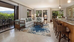 A spacious living area with a sofa, coffee table, and bar seating. Large windows offer a view of greenery.