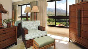 Amplia habitación con un ventanal que ofrece vistas a las montañas. Hay un sofá de mimbre con un cojín estampado, un armario de madera y una pequeña mesa con una lámpara.