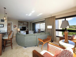 A hotel room with a sofa, chairs, a dining area, a TV, and a view of buildings.