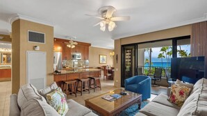 A modern living room with a sofa, coffee table, and a TV. There is a kitchen area with a bar and a view of the ocean. - Soothing Ocean Sounds and Views (Lahaina)