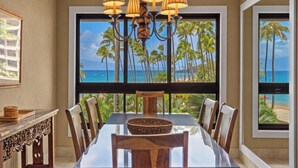 A dining area with a view of palm trees and the ocean, a chandelier, and a table with chairs. - Soothing Ocean Sounds and Views (Lahaina)