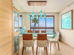 Un comedor con mesa de vidrio, sillas de madera, vista al océano y un cuadro en la pared.