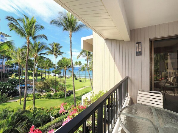 A balcony with a glass table and chairs, overlooking a landscaped garden with palm trees and a view of the ocean.