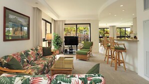 A living room with a sofa, chairs, a TV, and a dining area with a bar. - Iconic Ocean & Mountain Views | Corner Residence w/ Resort Amenities (Lahaina)