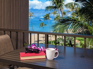 A table with a book, glasses, and a mug, overlooking a beach with palm trees and boats.