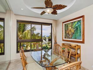 A dining area with a glass table, wicker chairs, a ceiling fan, and a view of palm trees.