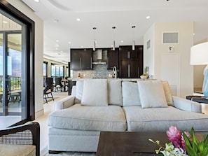 Una sala de estar moderna con un sofá gris, una zona de comedor con sillas, una cocina con armarios oscuros y lámparas colgantes.